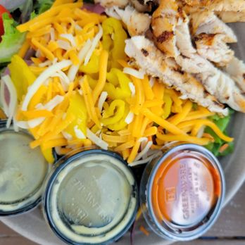 Thirsty Beaver - Salad - A plate of salad includes grilled chicken slices, shredded cheese, and mixed greens with yellow and purple vegetables. Three small containers of dressing are placed on the side, including ranch, honey mustard, and a red sauce. The dish appears to be served in a casual dining setting, emphasizing fresh ingredients and variety. - Burgers Near Me - Smithfield, Rhode Island