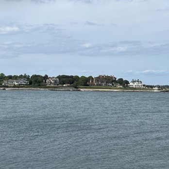 Cliff Walk - The image depicts a coastal landscape with a body of water in the foreground and a shoreline featuring various houses and large buildings. The structures are situated on a slightly elevated landmass, with some greenery visible around them. The sky is partly cloudy, and the scene appears calm and quiet, typical of a seaside residential area. - Parks Near Me - Newport, Rhode Island
