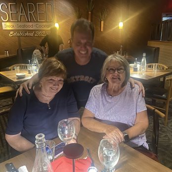 Seared - Team Wray. - A man is standing behind two elderly women who are seated at a wooden table in a restaurant. The women are smiling and appear to be enjoying their time. The table has glasses of water, a wine glass, and some bottles. The restaurant interior features wooden walls, ambient lighting, and a sign that reads 'SEARED' with additional text about steak, seafood, and cocktails. The setting suggests a casual dining experience. - Steakhouses Near Me - Toledo, Ohio
