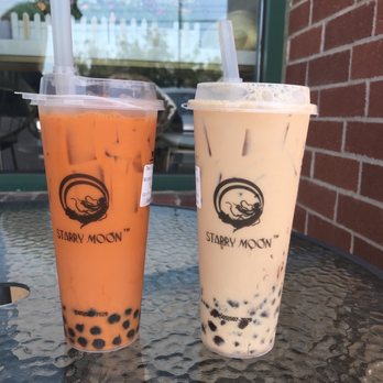 Starry Moon - Thai tea with boba and red bean milk tea - Two bubble tea drinks with tapioca pearls are positioned on a textured glass table outside a cafe. The left drink is orange-colored, and the right drink is a creamy beige. Both drinks are in clear cups with the 'Starry Moon' logo, and each has a wide straw. The background shows a brick wall, a window, and a blurred outdoor scene with a fence and some buildings. - Bubble Tea Near Me - Albuquerque, New Mexico