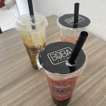 Boba Shack - Three boba drinks with black straws are arranged on a light wooden table inside Boba Shack. The drinks are in clear cups with the Boba Shack logo, and each has a plastic lid. The background shows a white wall and part of the shop's interior, indicating a casual dining environment focused on bubble tea beverages. - Bubble Tea Near Me - Apache Junction, Arizona