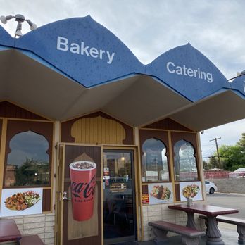 Caravan Grill - The image shows the exterior of a bakery and catering business with a blue awning displaying the words "Bakery" and "Catering." The storefront features large windows with images of food dishes and a door with a Coca-Cola advertisement. There are two concrete benches with red tops and a small table with a flower arrangement outside. The sky is partly cloudy, and a street with a parked car and some trees is visible in the background. - Halal Near Me - Tucson, Arizona