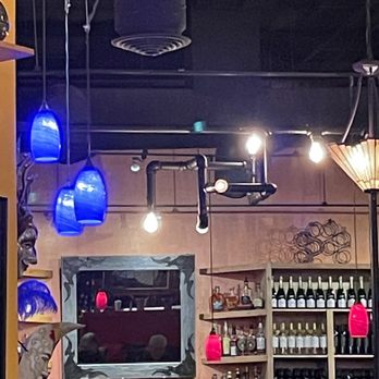 M'tucci's Italian - View of the bar - The image shows the interior of a bar with various lighting fixtures, including blue and pink hanging lights. Shelves filled with bottles of alcohol are visible on the right side, and a mirror is mounted on the wall behind the bar. The ceiling has exposed pipes and ductwork, contributing to an industrial aesthetic. A person is seen at the bar, and the overall setting suggests a lively, modern bar environment. - Italian Near Me - Albuquerque, New Mexico
