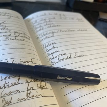 Penny Post - Kaweco Sport fountain pen in dark blue, and journal with Kaweco Caramel Brown ink for an idea list - A dark blue Kaweco Sport fountain pen is placed on an open notebook with handwritten notes, with a blurred background including office supplies. - Cards & Stationery Near Me - Alexandria, Virginia