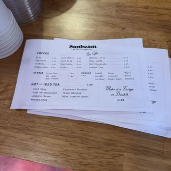 Sunbeam Bagel - drink menu - A printed menu from Sunbeam Bagel & Coffee Co. shows various coffee, tea, and syrup options on a wooden table with stacked plates and a binder in the background. - Coffee & Tea Near Me - Bryan, Texas