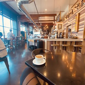 Coffee Plant Roaster - Sip, relax, enjoy - Inside a coffee shop with large windows, a coffee cup on a dark table is visible, with customers and baristas in the background. - Coffee & Tea Near Me - Eugene, Oregon