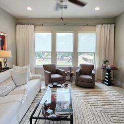 Budget Blinds of Southeast Austin and Hays County - Woven wood roller shades in the window diffuse the light & flowy white drapery panels add dimension & texture to frame the room in Driftwood