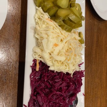 Grill Point - A rectangular white plate displays three sections of pickled vegetables on a wooden table. The top section has sliced cucumbers, the middle section contains shredded cabbage, and the bottom section features shredded red cabbage. A metal spoon is placed in the red cabbage section. The presentation is simple and typical of side dishes served in Middle Eastern or Eastern European cuisine. - Middle Eastern Near Me - Flushing, New York
