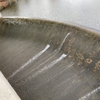 Taylor Park - Concrete dam structure with water flowing over the top edge into a body of water. The dam has a curved shape and appears to be part of a water management system. The water surface behind the dam is calm, and the surrounding area includes some natural elements like rocks and trees. - Parks Near Me - Millburn, New Jersey