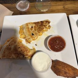 Chicken Quesadillas - A square white plate contains two chicken quesadillas, one with a browned, slightly charred surface, a fried fish fillet, a small cup of salsa, and a cup of sour cream. The plate is placed on a wooden table, with two small clear plastic cups and a napkin visible in the background.
