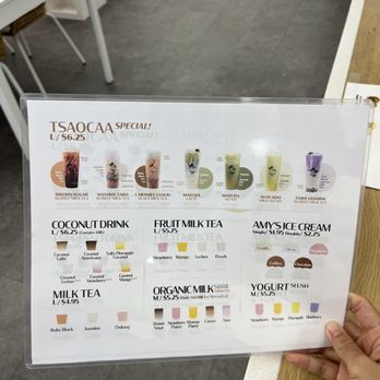Bubble + Tea By Tsaocaa - The menu board features a selection of bubble tea, milk tea, fruit milk tea, organic milk swirls, yogurt slush, and ice cream options, with prices ranging from $1.95 to $6.25. It includes flavor options such as brown sugar, matcha, avocado, taro, and more, with visual color indicators for each flavor. The menu is held in a transparent frame and is being held by a person in a casual setting, likely inside the bubble tea shop. - Bubble Tea Near Me - Austin, Texas