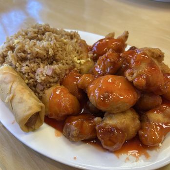 Royal Dragon - LL3. Sweet & Sour Chicken lunch option - A white oval plate holds sweet and sour chicken coated in a glossy orange sauce, alongside fried rice with small pieces of ham and scrambled egg, and a spring roll. The dish is set on a light-colored wooden table, with another plate of food visible in the background. - Chinese Near Me - Roy, Utah