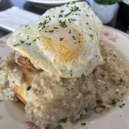 Chicken & Biscuits - A plate contains biscuits covered in gravy with visible chunks of meat and spices. A fried egg with a runny yolk is placed on top, garnished with chopped herbs. The dish is served on a decorative plate, with a restaurant setting visible in the background, including a napkin and some condiments.
