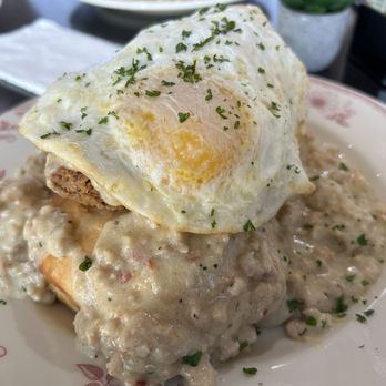 Eggstasy - Chicken & Biscuits - A plate contains biscuits covered in gravy with visible chunks of meat and spices. A fried egg with a runny yolk is placed on top, garnished with chopped herbs. The dish is served on a decorative plate, with a restaurant setting visible in the background, including a napkin and some condiments. - Breakfast & Brunch Near Me - Phoenix, Arizona