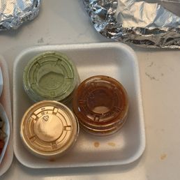 Sauces - Three small condiment containers with sauces, including green, brown, and reddish sauces, are placed on a foam tray on a white surface.