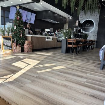 Kaffa Cafe - The photo depicts the interior of Kaffa Cafe, featuring a decorated Christmas tree with presents underneath near the counter. The cafe has a modern design with black and white walls, hanging greenery, and wooden flooring. Digital menu screens are visible behind the counter, and staff are working behind it. There are tables and chairs for customers, and the overall atmosphere is festive and contemporary. - Cafes Near Me - Crown Point, Indiana