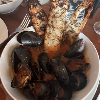 Sogno Restaurant - Mussels - A white bowl contains cooked mussels in a tomato-based broth, with some shells open revealing the meat. Three slices of toasted bread, topped with herbs and garlic, are placed upright in the bowl. The setting includes glasses of water, a fork, and other tableware, indicating a dining environment. - Wine Bars Near Me - St. Augustine, Florida