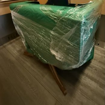 A2B Movers - Wrapped and padded the dining table and moved it with great care. - A piece of furniture, covered in green material and wrapped in clear plastic, is positioned on a wooden floor. The furniture appears to be on a small wooden stand or sled, and the setting suggests an indoor environment, possibly during a moving or packing process. - Movers Near Me - San Jose, California