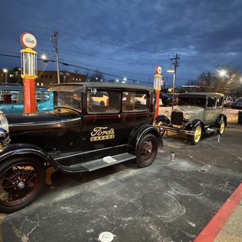 Ford's Garage Florence - Two vintage Ford automobiles are parked in a lot at Ford's Garage Florence, with illuminated vintage gas pump replicas nearby and a cloudy evening sky overhead. - Burgers Near Me - Florence, Kentucky