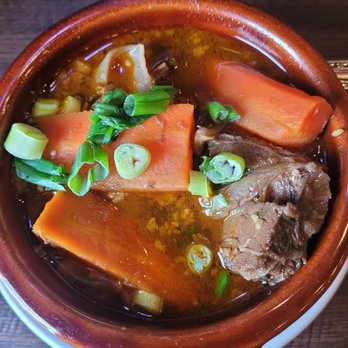 Pho 60 - Beef stew - A ceramic bowl filled with beef stew features large pieces of beef, thick carrot slices, and chopped green onions floating in a clear, savory broth. The bowl is set on a wooden table, with a glass of water visible in the background. The dish appears to be a traditional Asian-style beef stew with fresh garnishes. - Vietnamese Near Me - Brooklyn, New York