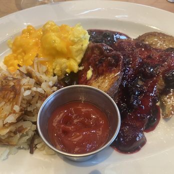 Jackson Square Grill - Wild Berry French toast - A breakfast plate includes French toast topped with a berry compote, scrambled eggs, and a portion of hash browns. A small metal cup of ketchup is placed on the plate. The dish is served on a white oval plate, with utensils visible in the background, indicating a dining setting at Jackson Square Grill. - Seafood Near Me - Columbus, Mississippi