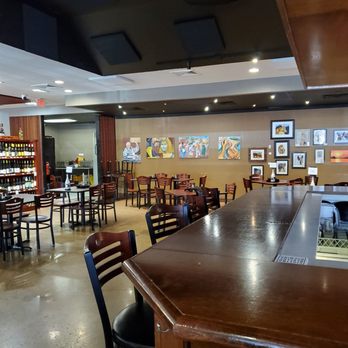 Ray's Wine Spirits Grill - From bar to part of dining area. You can see the beginning of the wine store half - A dining area with wooden chairs and tables, decorated with various artworks on the wall, and a bar counter in the foreground. - Wine Bars Near Me - Dayton, Ohio