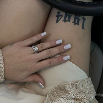 Apex Nail Spa - A hand with rings is placed on the person's thigh, which has a tattoo, inside a vehicle. The person has manicured nails and is wearing a beige knitted sweater. - Waxing Near Me - Montgomery, Illinois