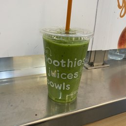 Apples and Greens - A green smoothie in a clear plastic cup with a domed lid and a brown straw is positioned on a metal counter. The cup has white text listing items like smoothies, juices, and bowls. The background includes a white wall and part of a menu or sign. The setting appears to be a juice or smoothie shop.
