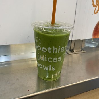 Jamba - Apples and Greens - A green smoothie in a clear plastic cup with a domed lid and a brown straw is positioned on a metal counter. The cup has white text listing items like smoothies, juices, and bowls. The background includes a white wall and part of a menu or sign. The setting appears to be a juice or smoothie shop. - Juice Bars & Smoothies Near Me - Chicago, Illinois
