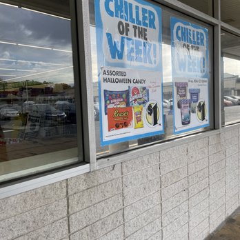 Dollar Tree - The image shows a storefront window with two promotional posters. The posters advertise Halloween candy and ice cream, with colorful images and pricing details. The window reflects the parking lot outside, which contains several parked cars and a cloudy sky. The building has a brick lower wall and a metal frame around the window. The interior lighting is visible through the glass. - Discount Store Near Me - Edison, New Jersey
