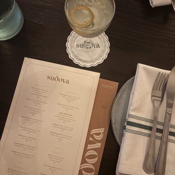 Sudova * Eastern European Kitchen - A cocktail glass with a lemon twist garnish sits on a napkin at a restaurant table, alongside a menu, water glass, and utensils. - Modern European Near Me - Cincinnati, Ohio