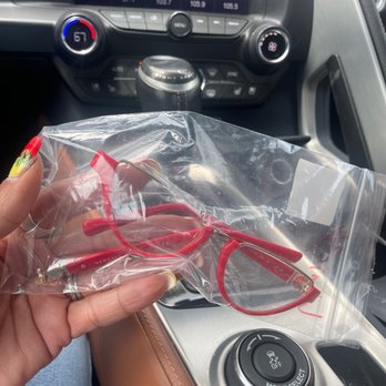 Schmidt's Optical and Hearing - My Versace glasses that was broken - A person with decorated nails holds a pair of red Versace glasses in a plastic bag inside a vehicle, with the car's dashboard and climate controls visible. - Eyewear & Opticians Near Me - Stuart, Florida
