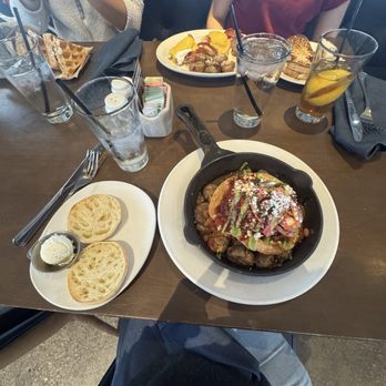 Hash Kitchen - Huevos Rancheros Hash * - The image shows a wooden table set with multiple breakfast dishes and beverages. There are two plates with food, one containing scrambled eggs, sausage, and potatoes, and another with a dish topped with sauce, cheese, and vegetables. Several glasses of water and iced tea with lemon are present, along with a small plate of sliced bread with butter. Two people are partially visible, seated at the table, with one wearing a beige sweater and the other in a red top. The setting appears to be a casual restaurant or brunch spot. - New American Near Me - Peoria, Arizona