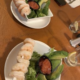 shrimp cocktail in question.... - Two plates of shrimp skewers are presented on a wooden table, each garnished with avocado slices, cucumber pieces, and a small container of salsa. The plates are white, and the setting includes a smartphone, a glass with lemon, and a napkin with cutlery in the background.