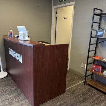 Ezdashcam - The image shows a reception desk made of dark wood with a computer monitor, hand sanitizer, and a small stand with a screen on top. To the left, there is a digital display stand with information. Behind the desk, a doorway leads to another room. To the right, a black metal shelving unit holds boxes, flags, and decorative items, including a small potted plant. The floor has a wood-like finish, and the walls are painted gray. - Auto Security Near Me - Tempe, Arizona