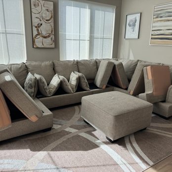 Pro Clean - A beige sectional sofa with multiple cushions and armrests is situated in a living room. The room features large windows with sheer curtains, allowing natural light to illuminate the space. The walls are decorated with abstract artwork, and a patterned area rug is placed underneath the sofa. The sofa appears to have some cushions removed or displaced, revealing the structure of the furniture. - Carpet Cleaning Near Me - Costa Mesa, California