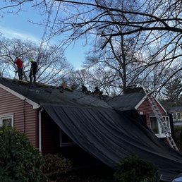 Two workers on the roof of a house are replacing or repairing shingles, with a ladder leaning against the house and a black tarp covering part of the roof.
