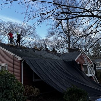 Pride Exteriors - Two workers on the roof of a house are replacing or repairing shingles, with a ladder leaning against the house and a black tarp covering part of the roof. - Roofing Near Me - North Attleboro, Massachusetts