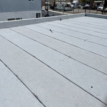 Capitol Roofing - A flat, white commercial roof with visible seams and a parapet wall along the edge. The roof surface appears to be made of a membrane material. In the background, there is a parking lot with several vehicles, a chain-link fence, and neighboring buildings. Utility poles and power lines are also visible in the distance. - Roofing Near Me - Commerce, California