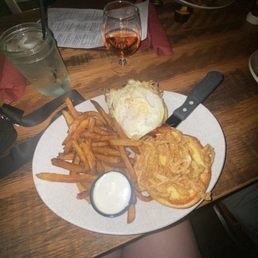 Hangover Burger - A plate of food includes a serving of French fries, a fried egg placed on a burger bun, and a large fried onion ring. A small container of white dipping sauce is also on the plate. The meal is set on a wooden table with a glass of rosé wine, a mason jar with a straw and water, and a menu in the background. The setting appears to be a restaurant or bar with dim lighting.