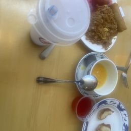 Good left by 2 hungry hungry people and it was so good!!!! Time to take this home! - The image shows a table with a variety of Asian dishes and beverages. There is a plate with fried rice, spring rolls, and skewered meat. A large drink with a straw is positioned at the top, and a bowl with a yellow liquid and a spoon is in the center. A small glass with red liquid and a plate with fried items are also visible. The table surface is light-colored wood, and utensils are placed around the dishes.