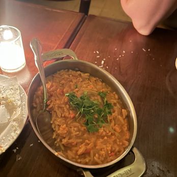 Swoony's - Creamy Lobster Orzo - A dish of lobster orzo with herbs is served in a metal bowl on a wooden table, with a spoon inside and a candle nearby. - New American Near Me - Brooklyn, New York