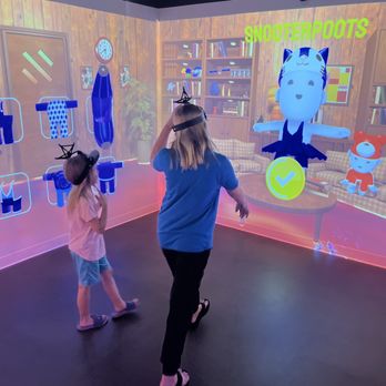 Immersive Gamebox - Kids playing floor is lava - Two children, one in a pink shirt and the other in a blue shirt, are standing in a dark room with a virtual reality game projected on the wall. The game features animated characters, including a large cat with a smiling face and a smaller robot, with colorful graphics and text. The children are wearing VR headsets and appear to be actively participating in the interactive experience. The room has a black floor and the projection displays a cozy, wood-paneled room with bookshelves and furniture, creating an immersive environment. - Party & Event Planning Near Me - Murray, Utah