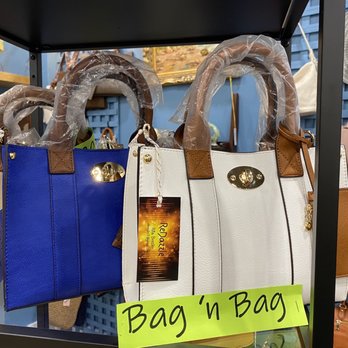 Painted Tree Kildeer - Picked up a nice bag - Several handbags in blue, white, and brown are arranged on a display shelf. The bags have gold hardware and are wrapped in plastic handles for protection. A yellow sign labeled "Bag 'n Bag" is attached to the display, indicating the store's branding. The background includes additional accessories and decorative items, with a blue wall and other display shelves visible. - Home Decor Near Me - Kildeer, Illinois