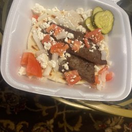 a meal in a plastic container - A foam takeout container holds a taco with a soft tortilla base topped with cooked beef, chopped tomatoes, onions, shredded cheese, and sliced pickles. The ingredients are arranged in a casual presentation, with some ingredients spilling over the edges of the tortilla. The background shows a patterned carpet, indicating the photo was taken indoors.