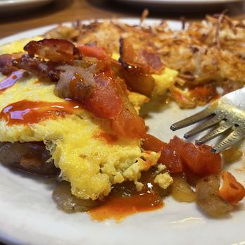 Denny's - A breakfast plate contains scrambled eggs topped with ketchup, crispy bacon strips, and a portion of hash browns. A fork is placed on the plate, and the food appears partially eaten. The plate is on a wooden table, with other plates visible in the background. - American Near Me - Raton, New Mexico