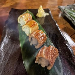 a plate of sushi on a leaf - Sushi rolls topped with eel and sauce are arranged on a green leaf on a black ceramic plate, with wasabi in the background.