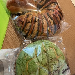 Red Bean Bun - Two baked buns are shown in clear plastic packaging on a wooden surface. The top bun has a golden-brown crust with black sesame seeds and a drizzle of dark syrup. The bottom bun is green with a cracked surface, indicating a matcha flavor, and has a similar drizzle. The packaging features a cartoon character and some text, suggesting they are from a bakery specializing in Asian-style baked goods.