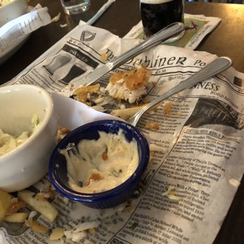 Two Fools Tavern - What's left of fish and chips - A plate with leftover fish and chips, including tartar sauce in a small blue dish, is placed on newspaper-style wrapping on a pub table. There are utensils, a glass of dark soda, and a pint of beer nearby. The setting appears to be a casual Irish pub with a wooden table and other food and drink items visible in the background. - Irish Near Me - Albuquerque, New Mexico