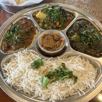 Curry Masala - Spring - Vegetarian Thali - A vegetarian Indian thali is presented on a metal platter with separate compartments. It includes a portion of white basmati rice garnished with cilantro, three different vegetable curries topped with fresh herbs, and a small container of chutney or pickle in the center. In the background, a basket of naan bread with herbs is partially visible. The setting suggests a restaurant serving authentic Indo-Pak cuisine with a variety of vegetarian options. - Indian Near Me - Spring, Texas